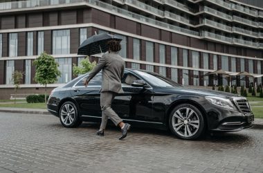 Man holding umbrella opens car door on rainy day in front of modern building.