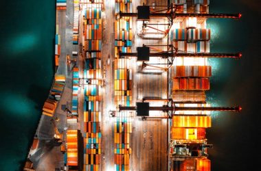 Dramatic aerial view of a busy shipping port with colorful containers and bright lights at night.